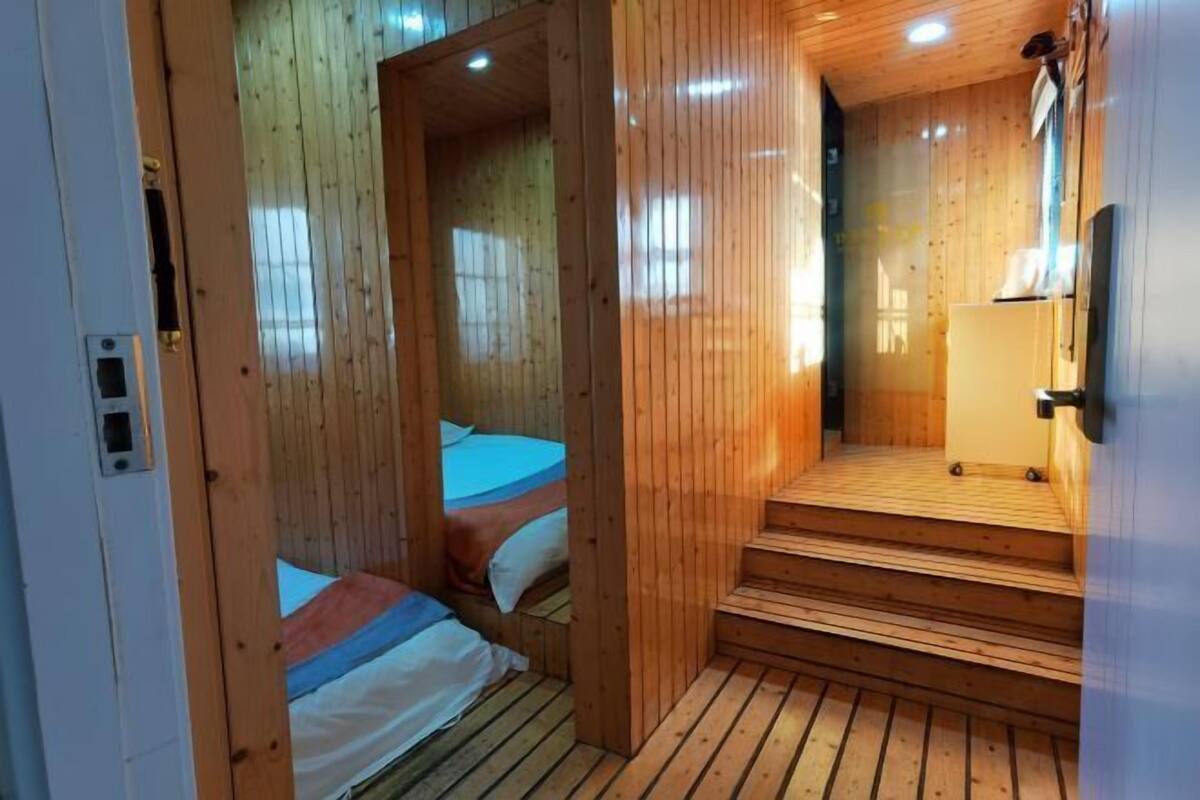 Une cabane en bois avec un petit escalier menant à une porte, un lit recouvert d’une couverture bleue et blanche, et un plancher en bois.