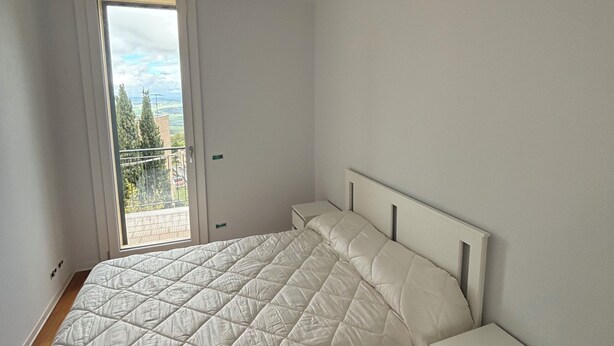 Un dormitorio con una cama grande, una mesita de noche y un ventanal con vistas a árboles y edificios.