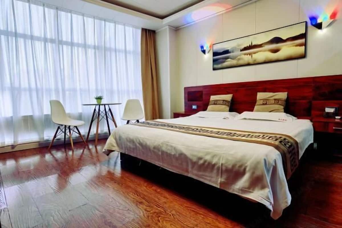A hotel room with a large bed, a desk with a chair, a window with curtains, and a painting on the wall.