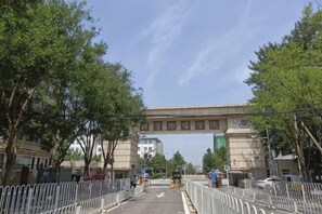 Una entrada con reja, un letrero, árboles y edificios al fondo.