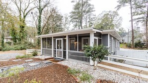 Ett litet hus med en inglasad veranda, omgivet av en trädgård med stenplattor och ett litet träd.