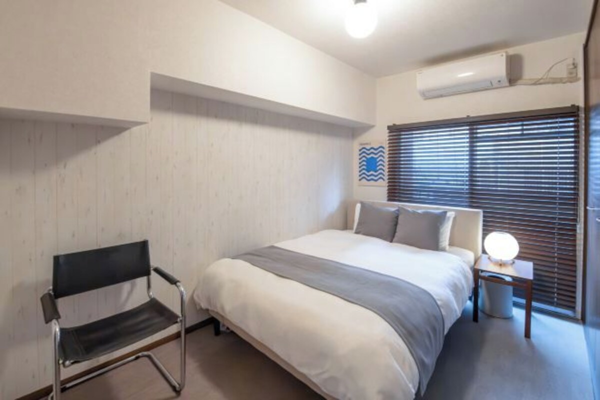 Een slaapkamer met een bed, een stoel, een nachtkastje met een lamp, een raam met jaloezieën en een airconditioning aan de muur.