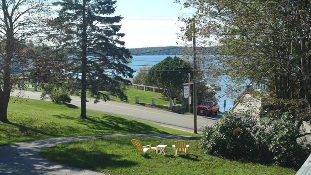 A grassy area with a bench and chairs facing a road and a body of water.
