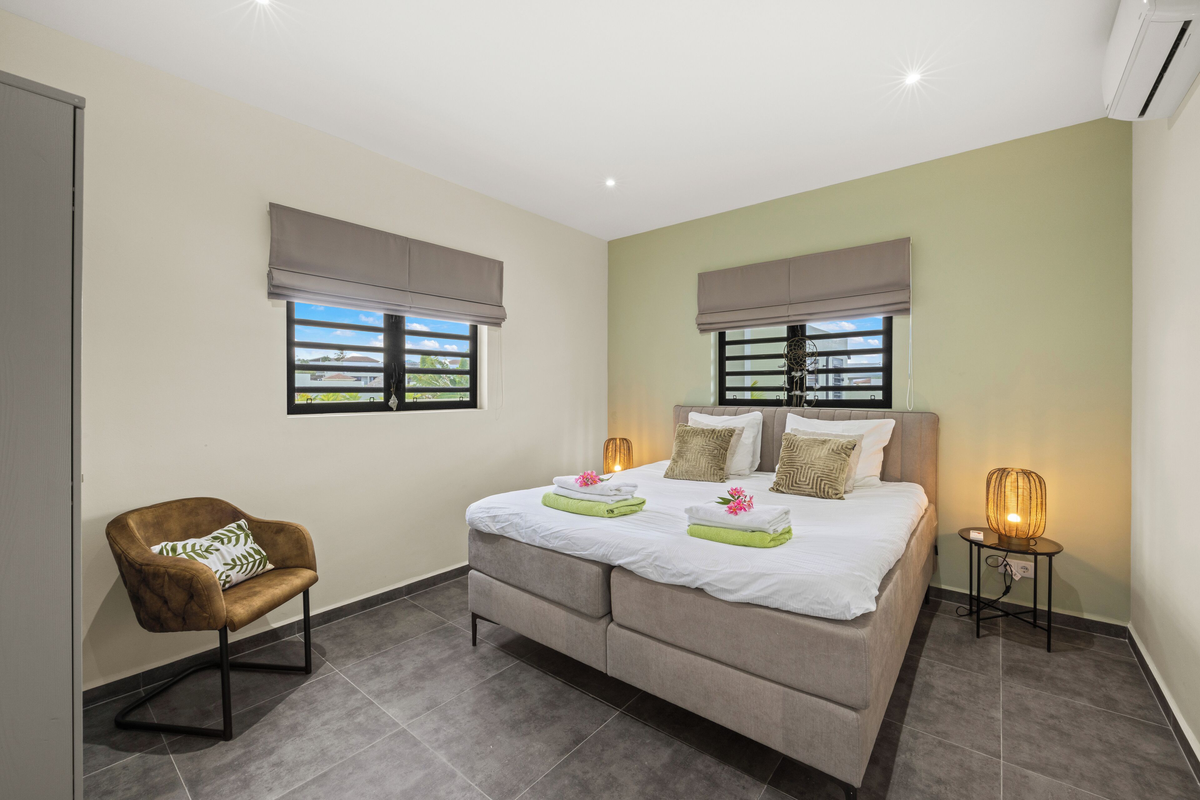 A bedroom with a bed, two bedside tables, a chair, and two windows with blinds.