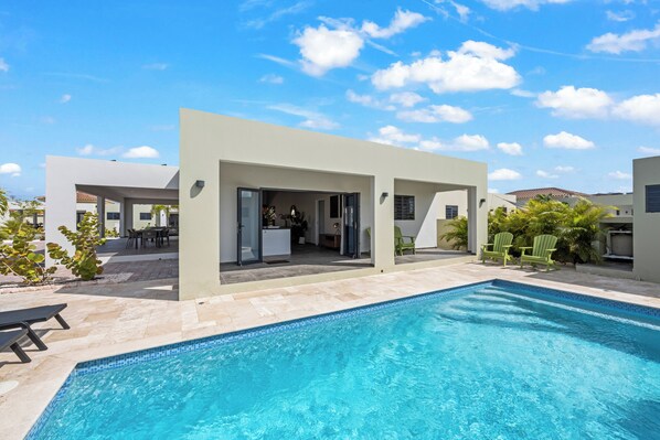 A modern pool area with a clear blue pool, a covered seating area, and a building with an open entrance. - Villa Amber with pool at mambo beach (Willemstad)