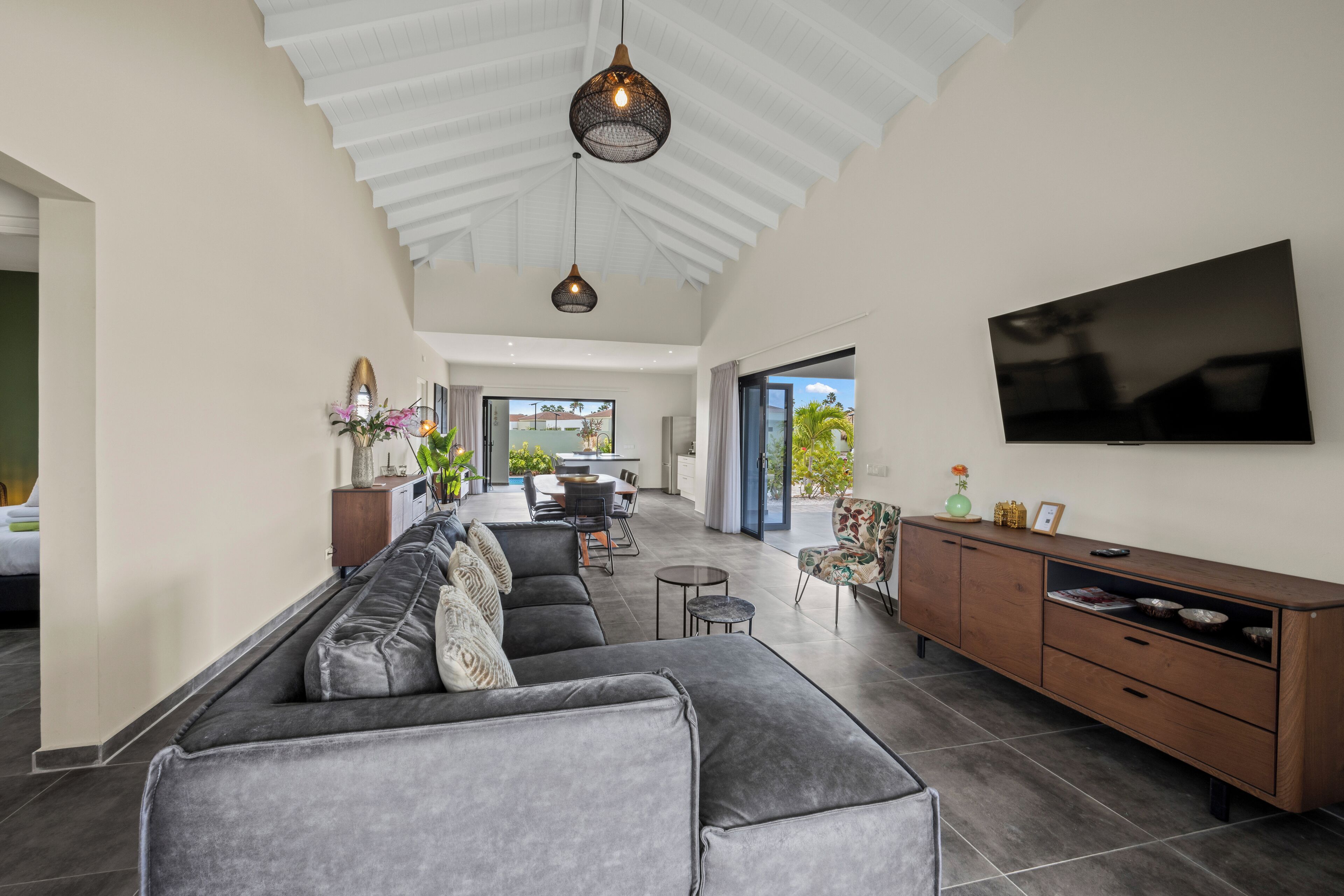A spacious living room with a grey sectional sofa, a wooden TV stand, and a large flat-screen TV mounted on the wall.