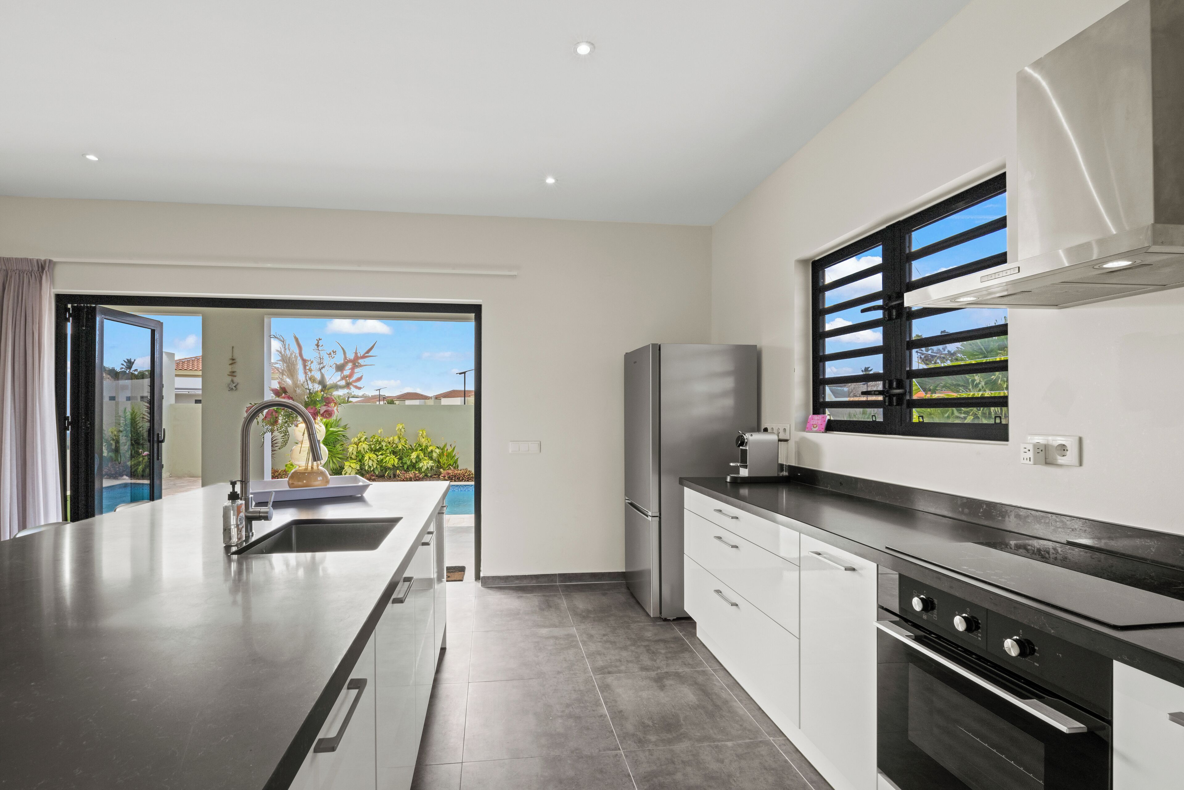 A modern kitchen with a stainless steel refrigerator, a built-in oven, and a large window with black blinds.