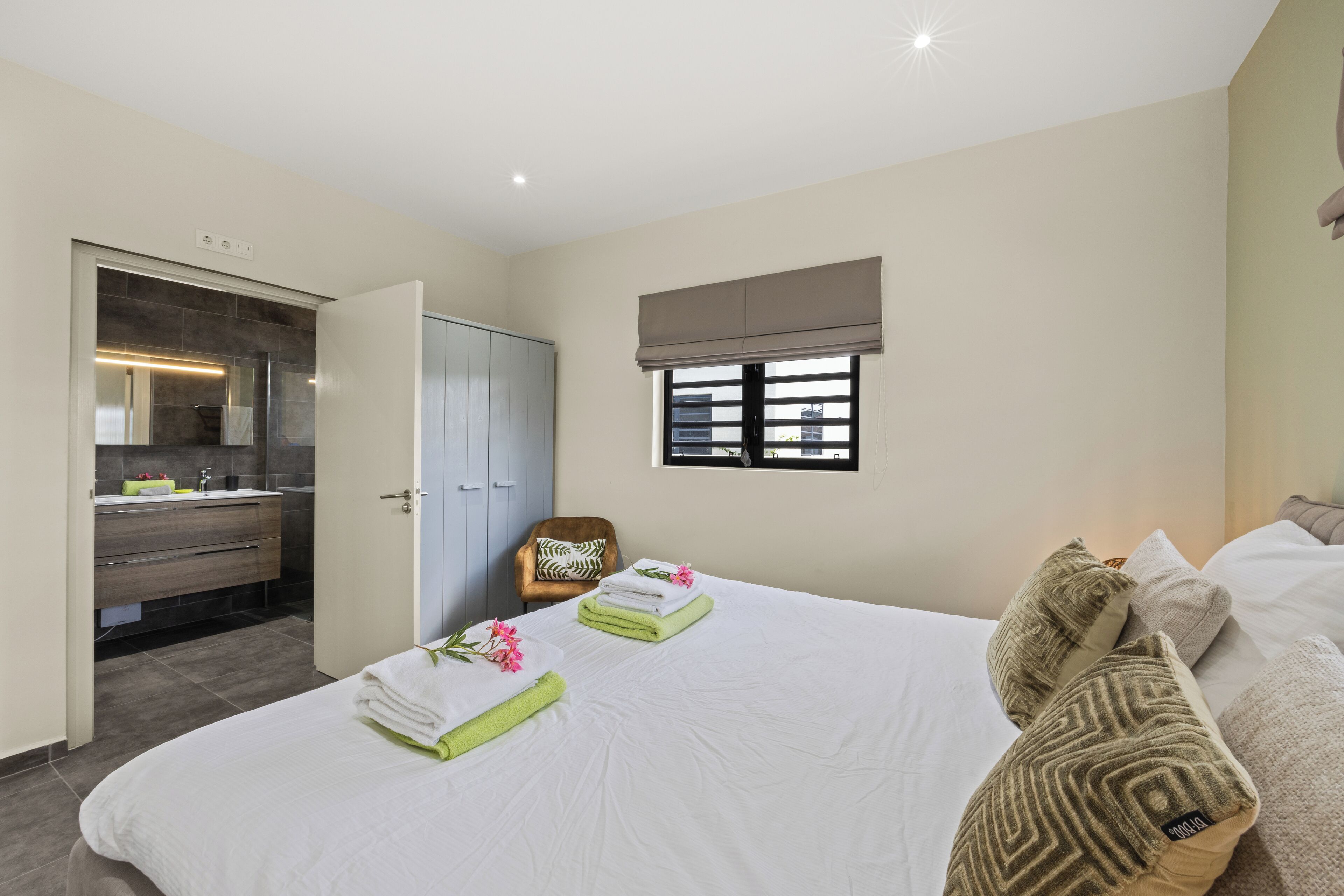A bedroom with a neatly made bed, a window with a view, a wardrobe, and a bathroom with a mirror and sink.