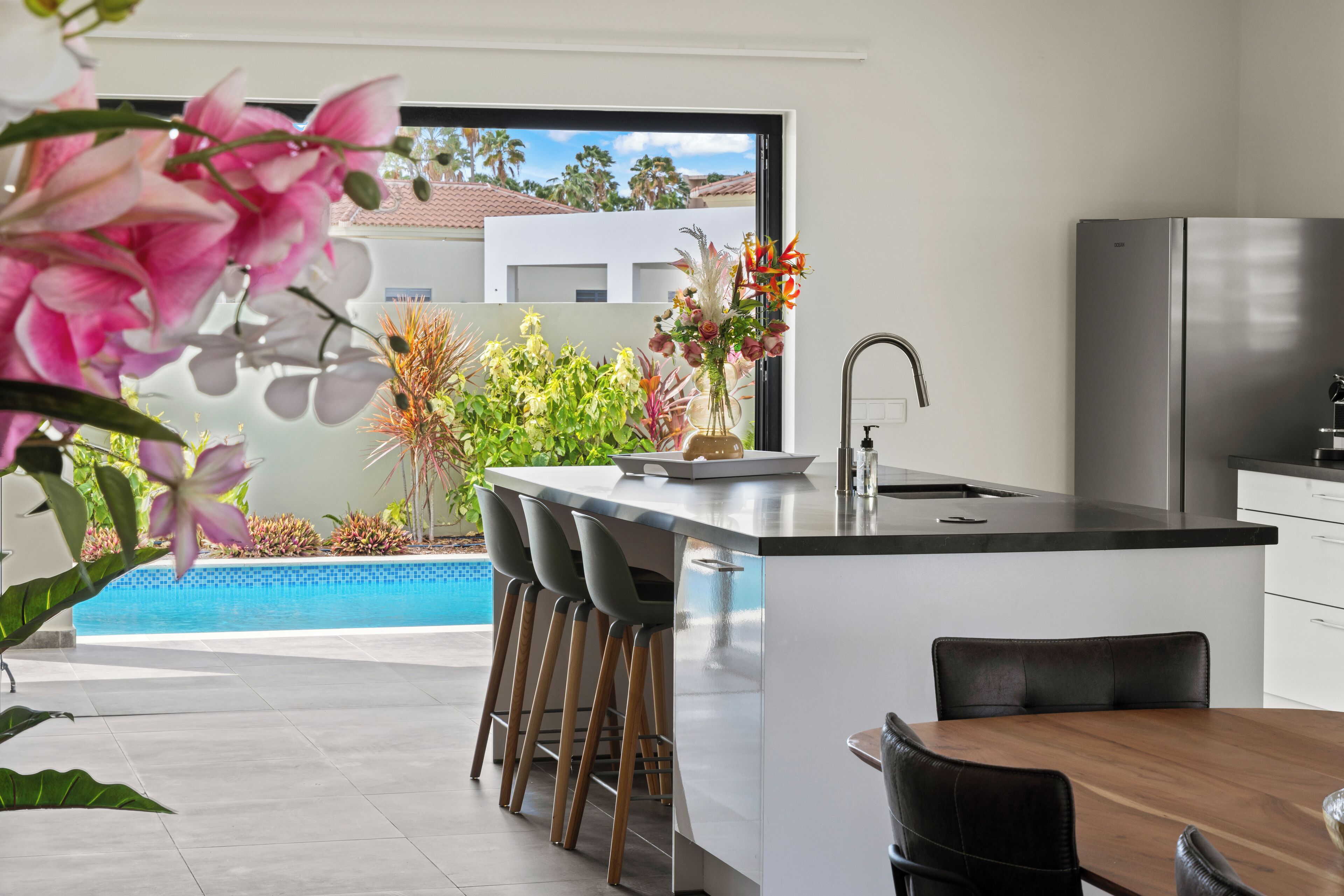 A modern kitchen with a large island, bar stools, a dining table, and a view of a swimming pool and greenery.