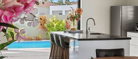A modern kitchen with a large island, bar stools, a dining table, and a view of a swimming pool and greenery.