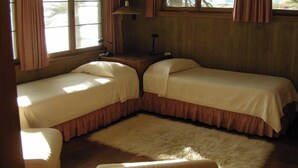 A room with two single beds, a small table, a lamp, and a window with curtains.