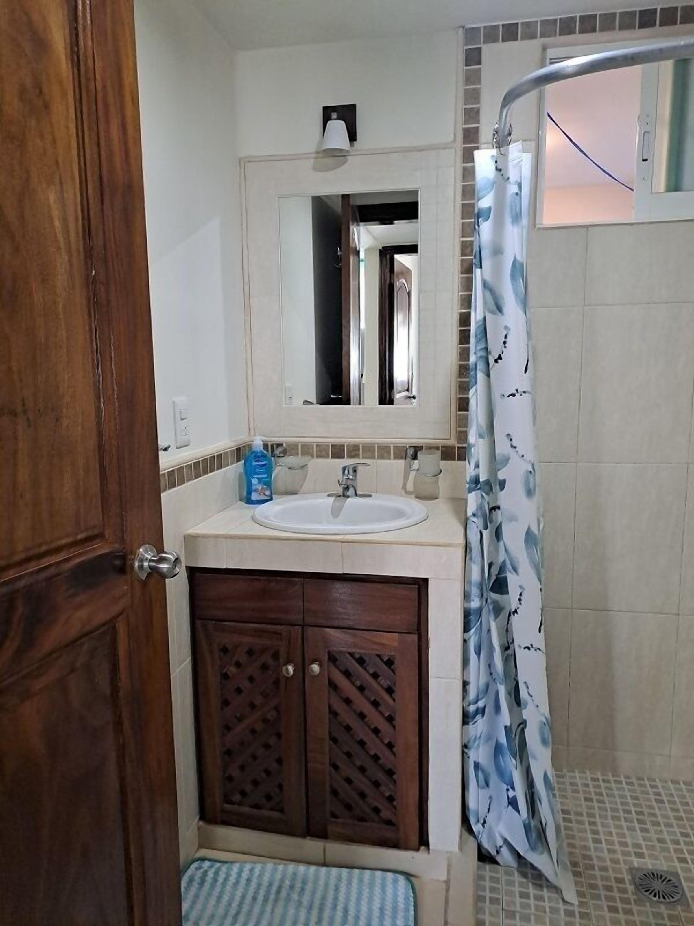 Un bagno con porta in legno, un lavandino con specchio sopra, tenda per la doccia e pavimento piastrellato.