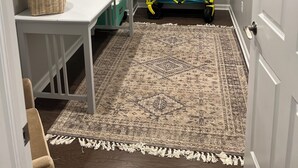 Una habitación compacta con un escritorio blanco, cestas de mimbre, un cochecito azul y una alfombra estampada.