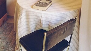 Una mesa redonda pequeña con mantel a rayas, un jarrón blanco con flores y un libro. Una silla está colocada frente a la mesa.