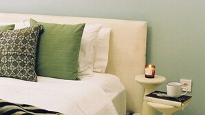 Un dormitorio con cama, almohadas, una mesita de noche con una vela y una pequeña mesa con una taza y un libro.