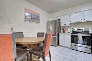 Cozinha compacta com armários brancos, geladeira de aço inoxidável, fogão e área de jantar com mesa redonda e cadeiras.