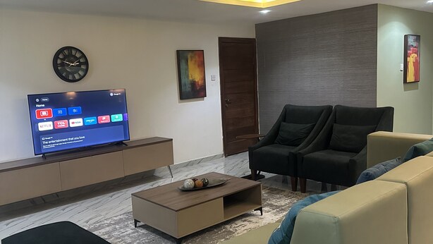 A modern living room with a flat-screen TV, a sofa, a coffee table, and a decorative clock. - Live, work,& chill in style—4BR Apartment in Central VI (Lagos)