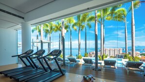 Une salle de sport équipée de tapis de course et d’un banc, avec vue sur une piscine bordée de chaises longues et de palmiers.