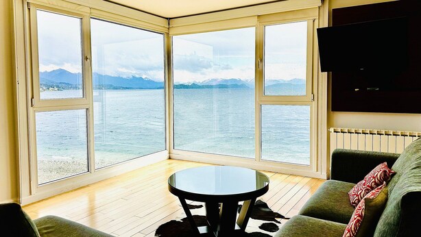 A corner room with a large bay window overlooking a body of water and mountains. There is a round table and a sofa with a patterned throw. - Oasis apartment on the Nahuel Huapi Lake (San Carlos de Bariloche)