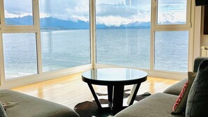 A corner room with a large window overlooking a body of water and mountains. There is a round table and two sofas. - Oasis apartment on the Nahuel Huapi Lake (San Carlos de Bariloche)