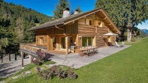 A wooden cabin with a deck, surrounded by a grassy area and trees. - Chalet, 200m from ski slopes, terrace, balcony, fireplace or stove, parking, tv, ski locker, 200m² (La Clusaz)
