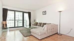 A modern living room with a glass coffee table, a beige sofa, a small ottoman, a floor lamp, and a view of a cityscape through large windows. - Heart of Canary Wharf. Stunning dock views. Close to all amenities. (London)