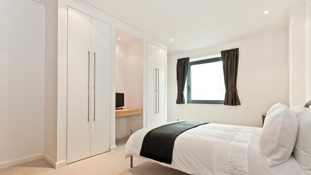A bedroom with a bed, a wardrobe, a desk with a computer, and a window with curtains. - Heart of Canary Wharf. Stunning dock views. Close to all amenities. (London)
