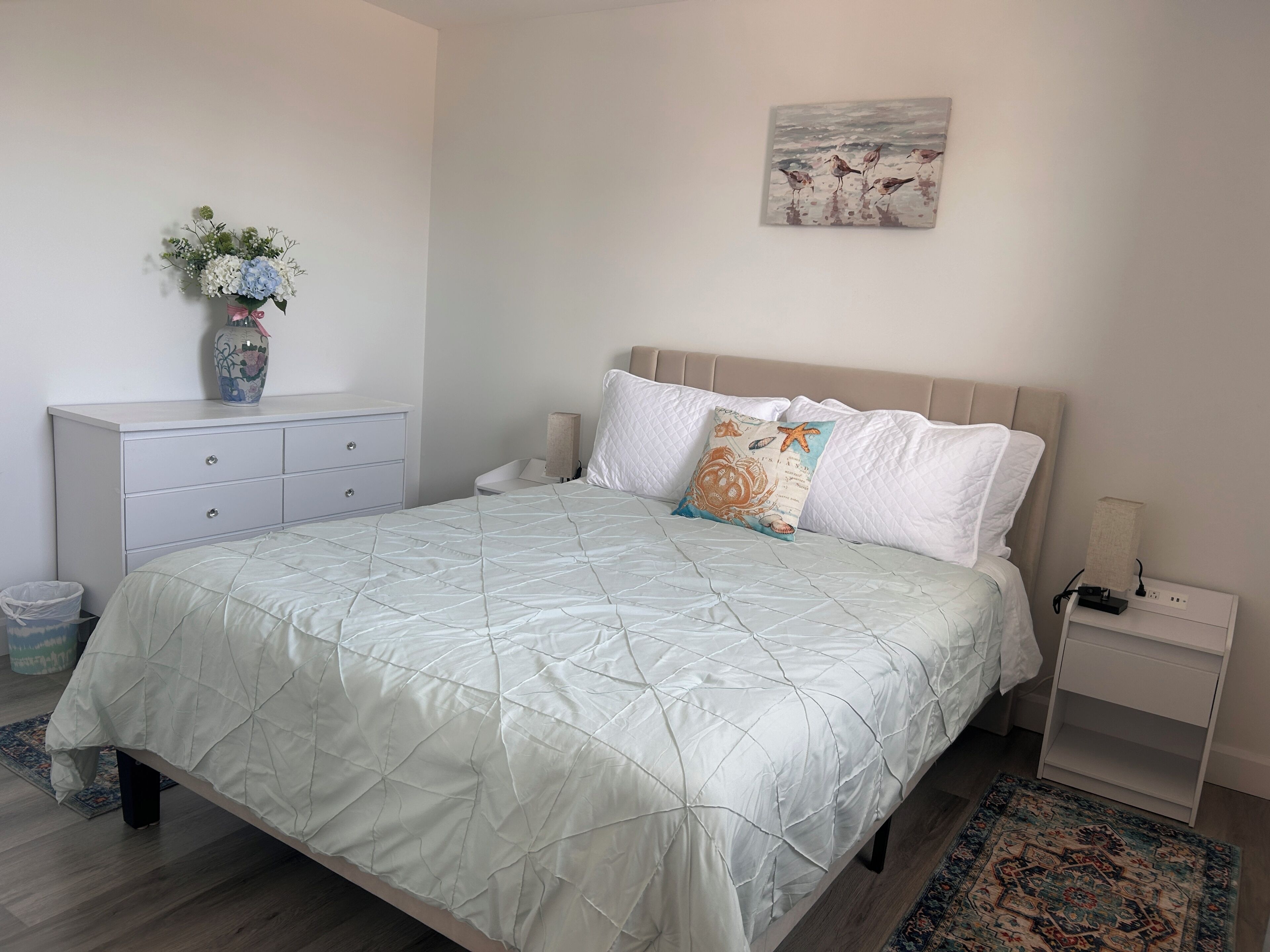 A bedroom with a bed, bedside tables, a nightstand, a vase with flowers, and a wall painting.