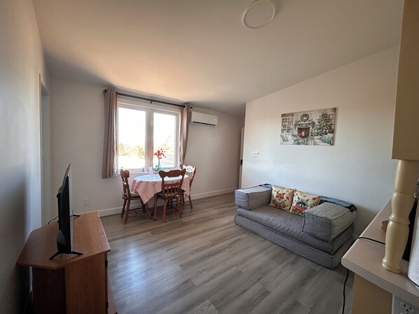 Ein Wohnzimmer mit einer Couch, einem Esstisch mit Stühlen, einem Fernseher auf einem Schrank und einem Gemälde einer Kaminfeuerstätte an der Wand.