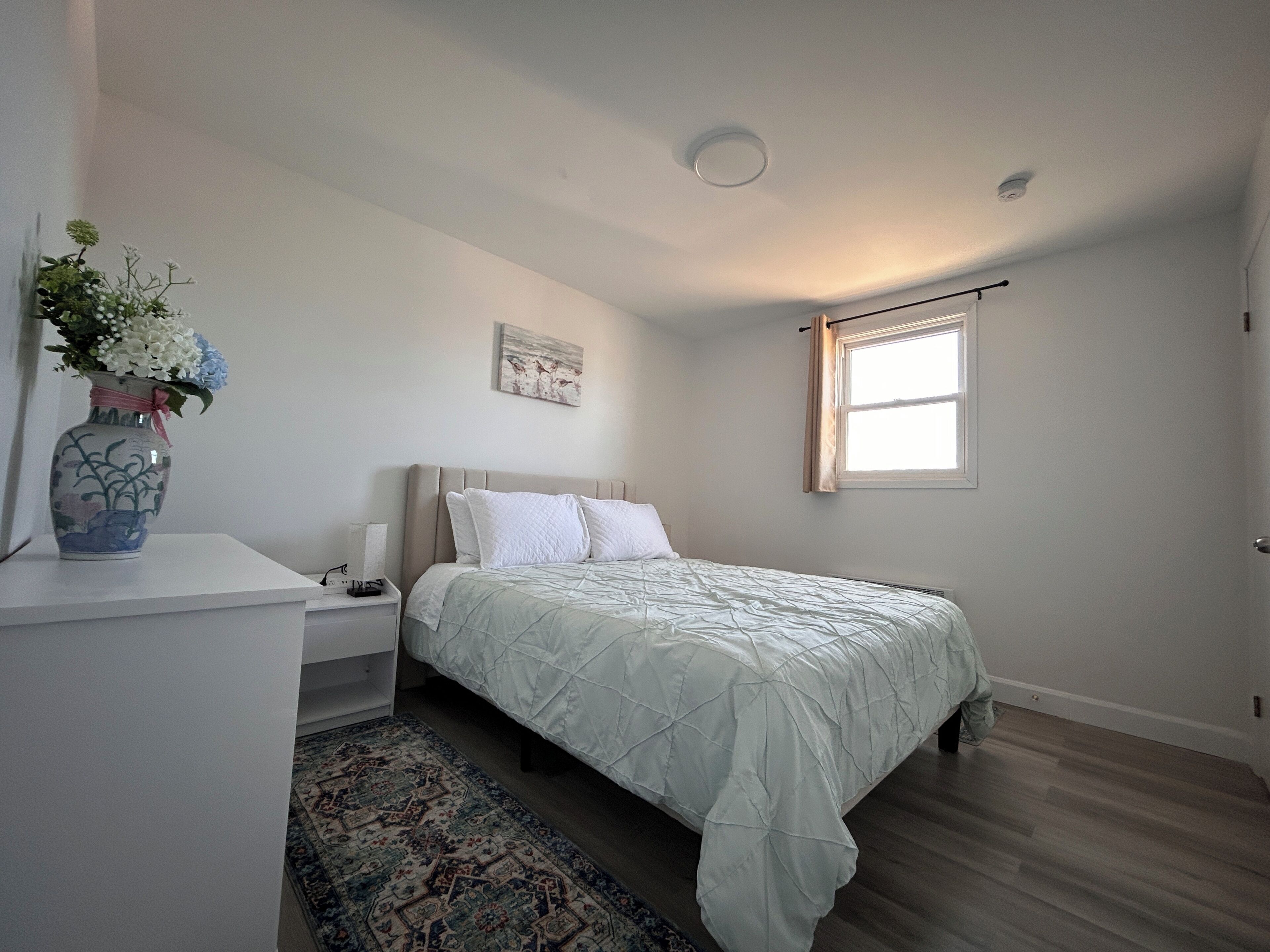 A bedroom with a bed, a nightstand, a vase with flowers, a rug, and a window with curtains.