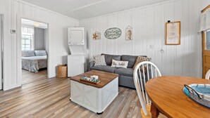 Una habitació amb sòl de fusta, un sofà gris, una taula de café blanca, una taula de menjador i una cadira blanca.
