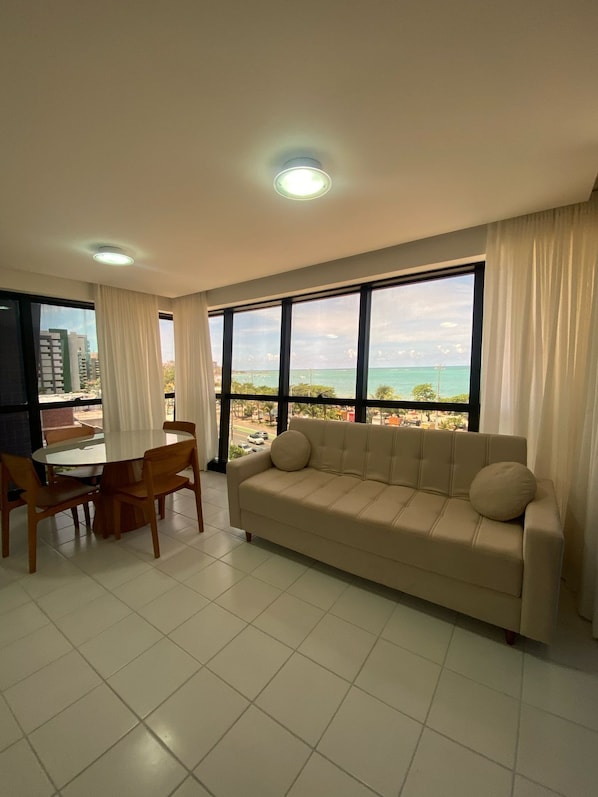 A living room with a sofa, dining table, chairs, and large windows with a view of the ocean and buildings. - Edificio Neo Apto 401-Mandi H (Maceio)