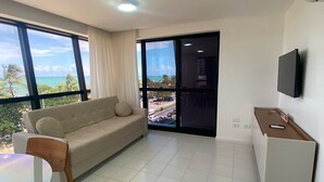 A modern living room with a sofa, a dining table, and chairs. There is a TV mounted on the wall and a balcony with a view of the ocean. - Edificio Neo Apto 401-Mandi H (Maceio)