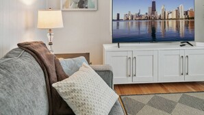 Ein Wohnzimmer mit einem großen Flachbildfernseher, der eine Stadtansicht zeigt, einem weißen Schrank, einem grauen Sofa und einem gemusterten Teppich.