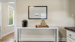 Een witte console tafel met een koffiezetapparaat, een dienblad met koffiekopjes en een ingelijst foto aan de muur.