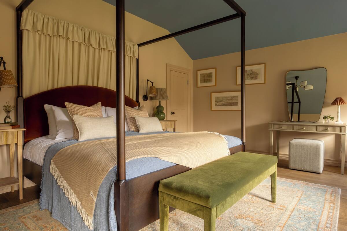 A four-poster canopy bed with a wooden headboard, a bench, and a bedside table with a lamp.