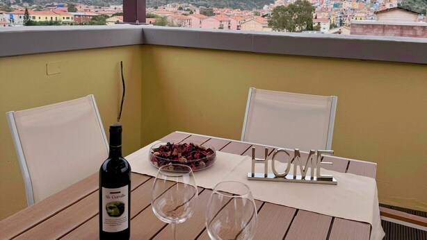 A balcony with a table set for two, a bottle of wine, and a bowl of grapes. The view includes a town and mountains in the background. - SkyView Apartment in BOSA (Bosa)