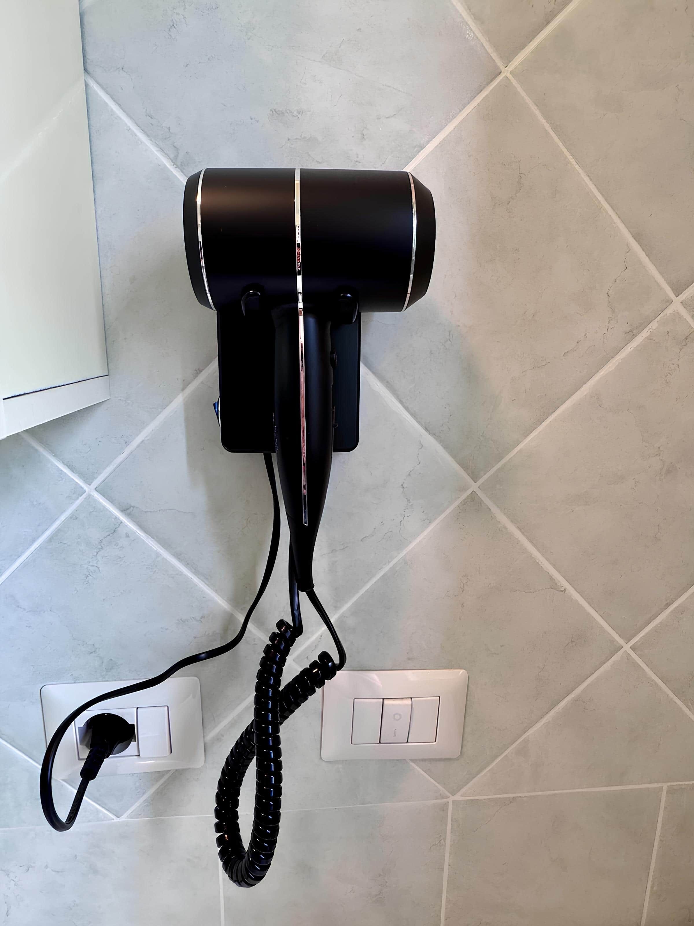 Um secador de cabelo fixado numa parede revestida de azulejos, com o cabo ligado a uma tomada.