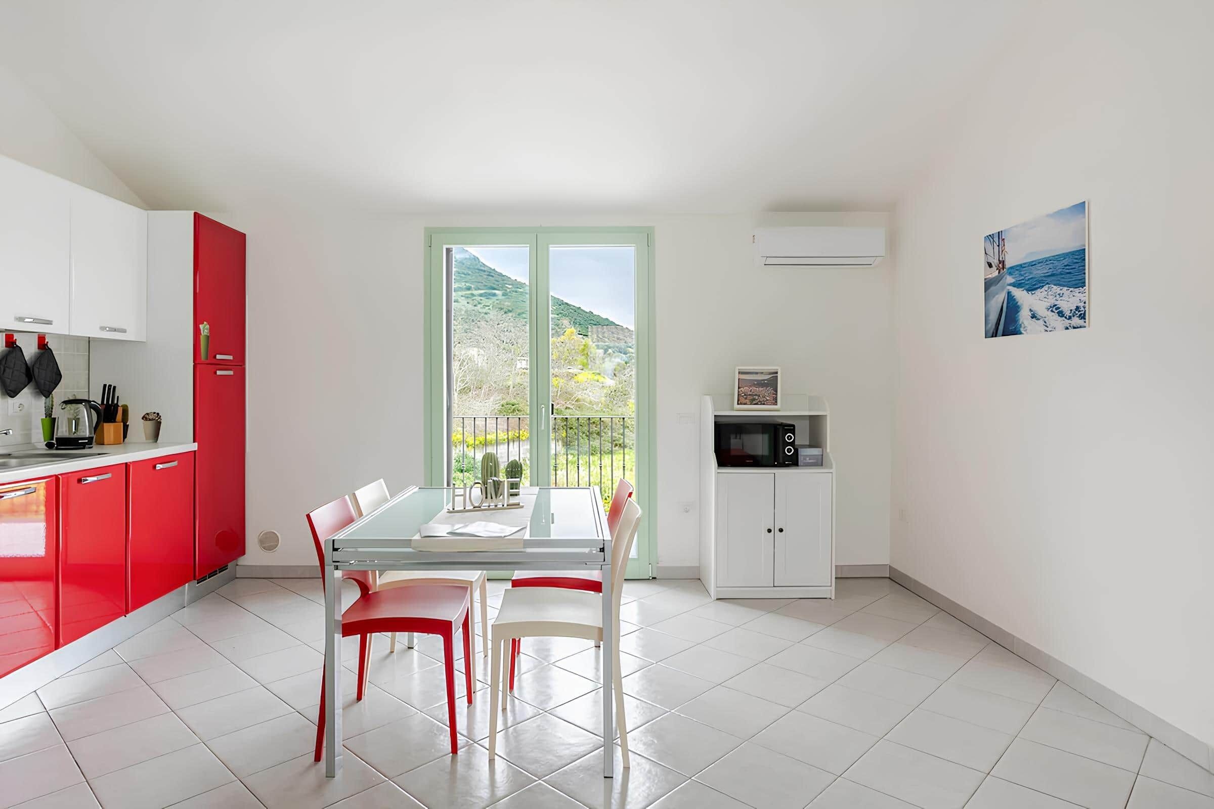 Une cuisine moderne avec des meubles rouges, une table à manger avec des chaises, une vue à travers des portes vitrées et un four à micro-ondes.