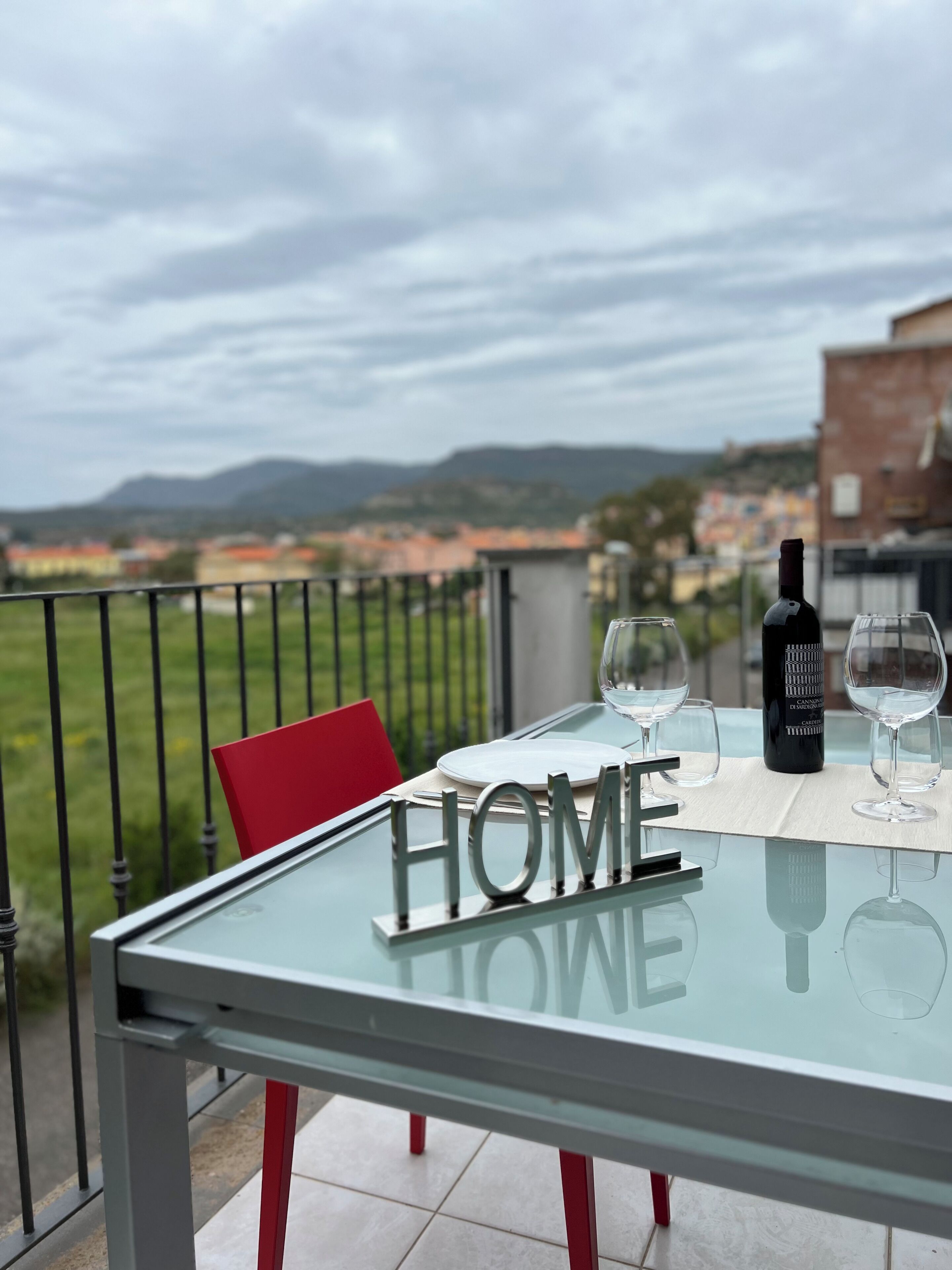 Ein Glastisch mit einem "HOME"-Schild, zwei Weingläsern und einer Weinflasche auf einem Balkon mit Blick auf eine Stadtlandschaft.