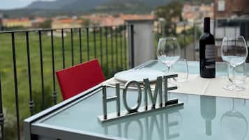 Ein Glastisch mit einem "HOME"-Schild, zwei Weingläsern und einer Weinflasche auf einem Balkon mit Blick auf eine Stadtlandschaft.