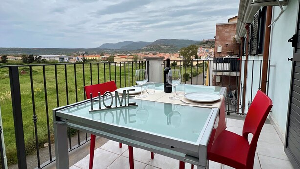 A balcony with a glass table and red chairs, overlooking a cityscape and mountains. - Sunset Apartment in BOSA (Bosa)