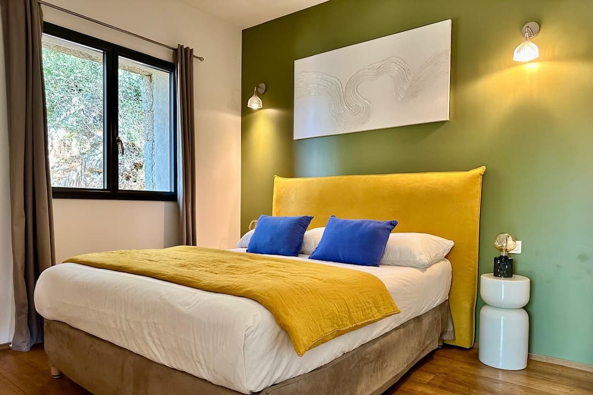 A modern bedroom with a large bed, a yellow headboard, and a view of trees through the window.
