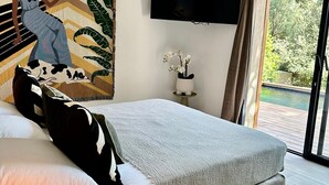 A modern bedroom with a large bed, a flat-screen TV, a window with a view of greenery, and a small table with a lamp. - Les Bergeries de Figari (Figari)