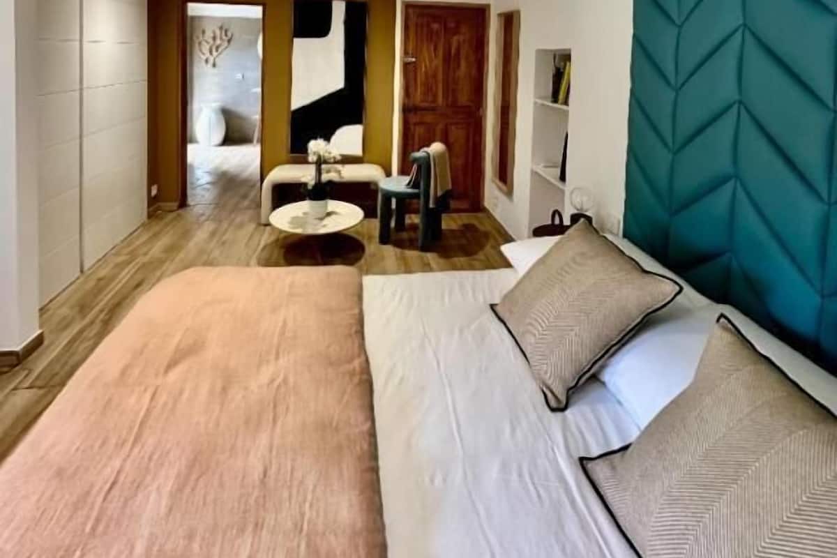 A modern hotel room with a large bed, a small sitting area, and a view into another room.