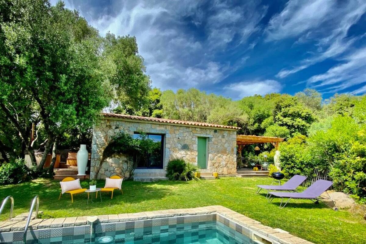 A swimming pool with a stone border, a stone building with a green door, and two lounge chairs.