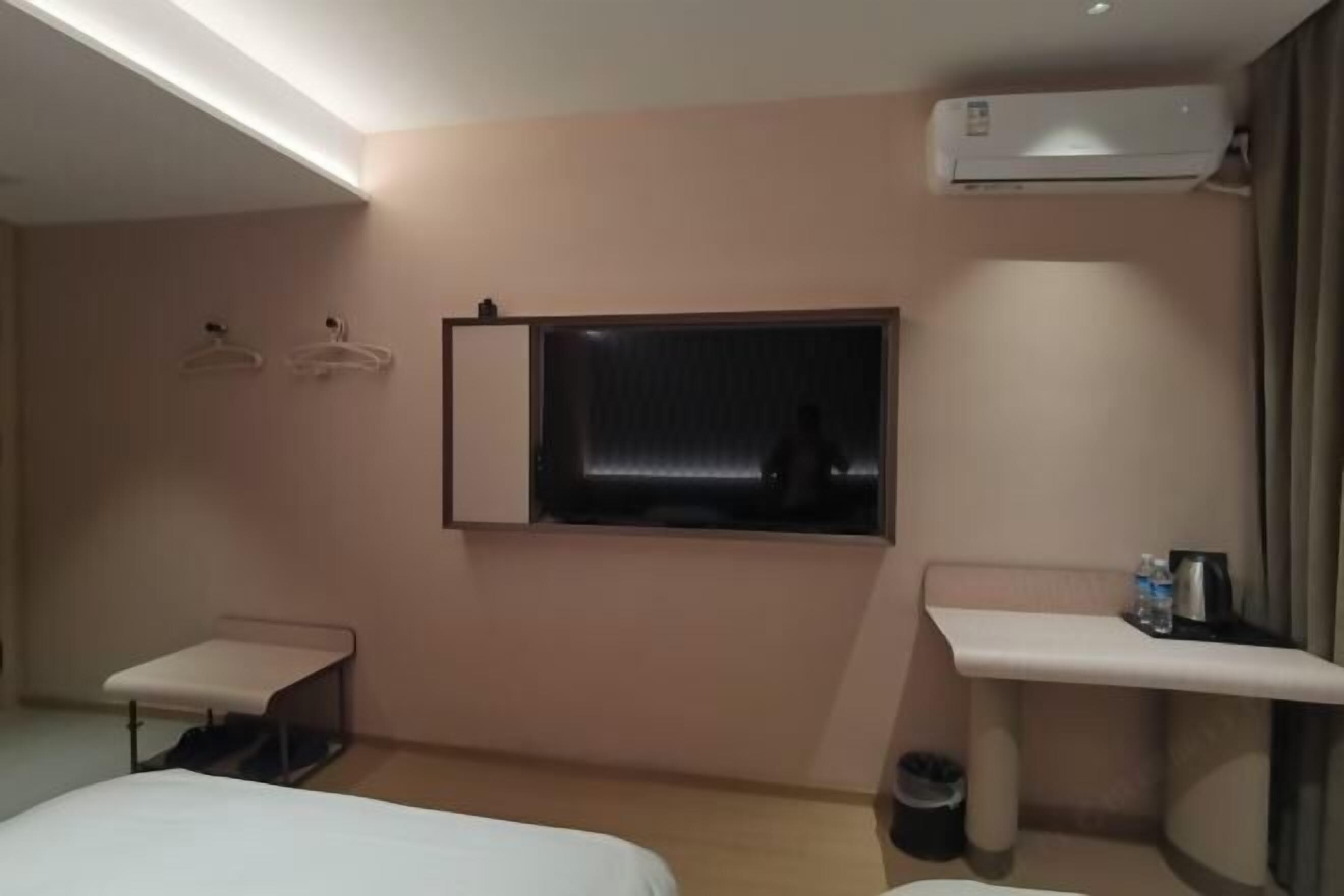 Habitación de hotel con cama, una mesita, ventana, equipo de aire acondicionado y un estante con tetera y botella de agua.