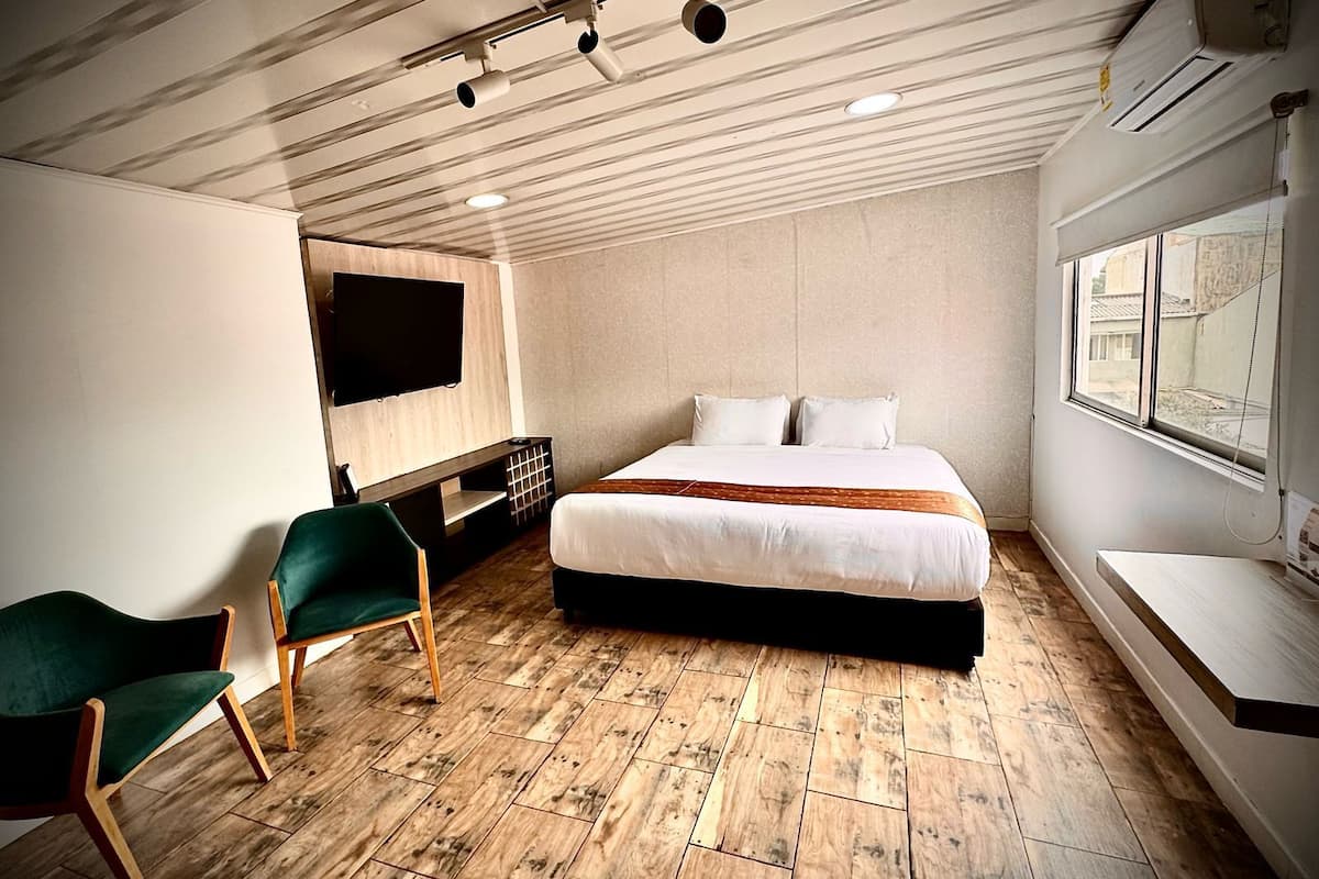 Habitación de hotel moderna con piso de madera, cama con ropa blanca, una silla verde, televisor de pantalla plana montado en la pared y ventana con vista.
