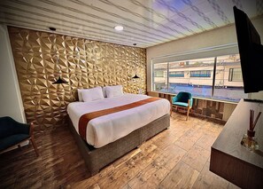 A modern hotel room with a geometric-patterned wall, a large bed, a seating area with a chair, and a view of the outside through a large window. - HOTEL AMERICAN VISA NORMANDIA  (Bogotá)