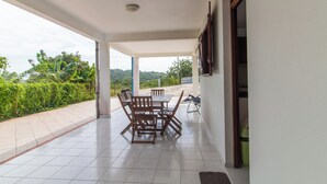 A covered patio with a dining table and chairs, a view of greenery and hills, and a tiled floor.